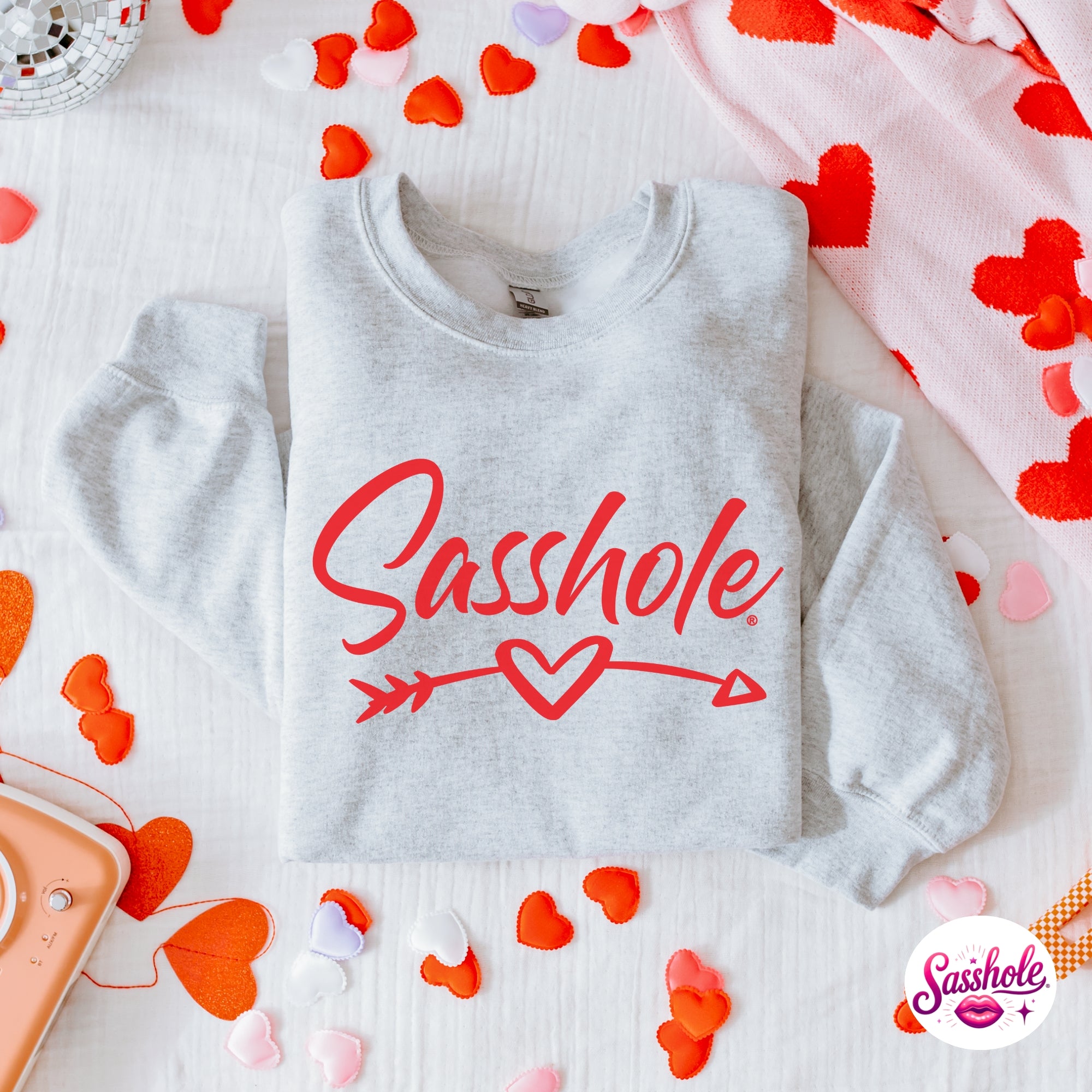 Image of Gray sweatshirt with 'Sasshole' text and heart design on a surface with heart-shaped confetti.