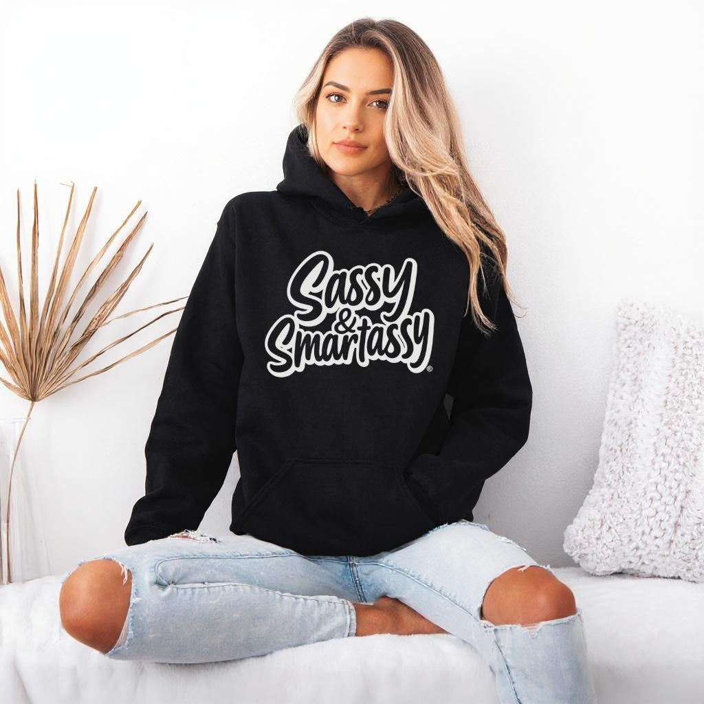 Woman wearing a black hoodie with 'Sassy & Smartassy' text, sitting on a white couch.