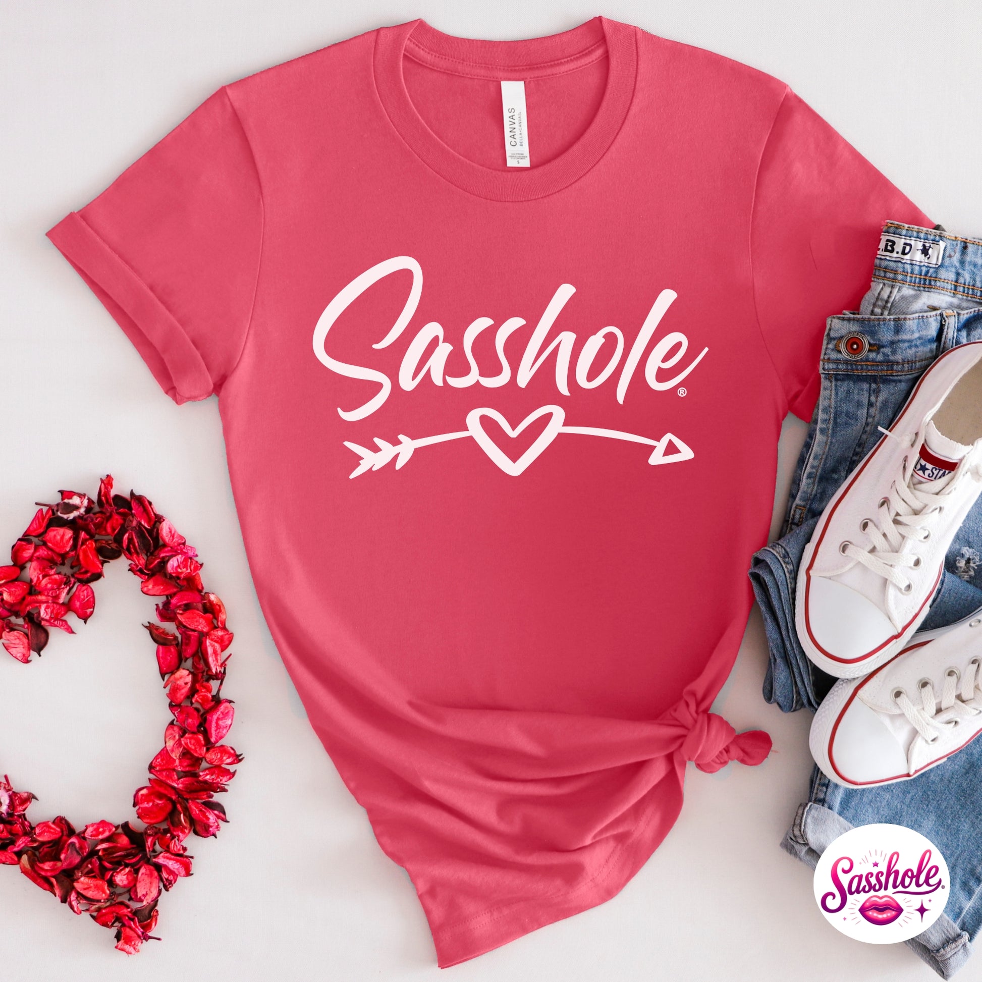 Pink t-shirt with 'Sasshole' graphic, surrounded by denim jeans and white sneakers on a light gray background.