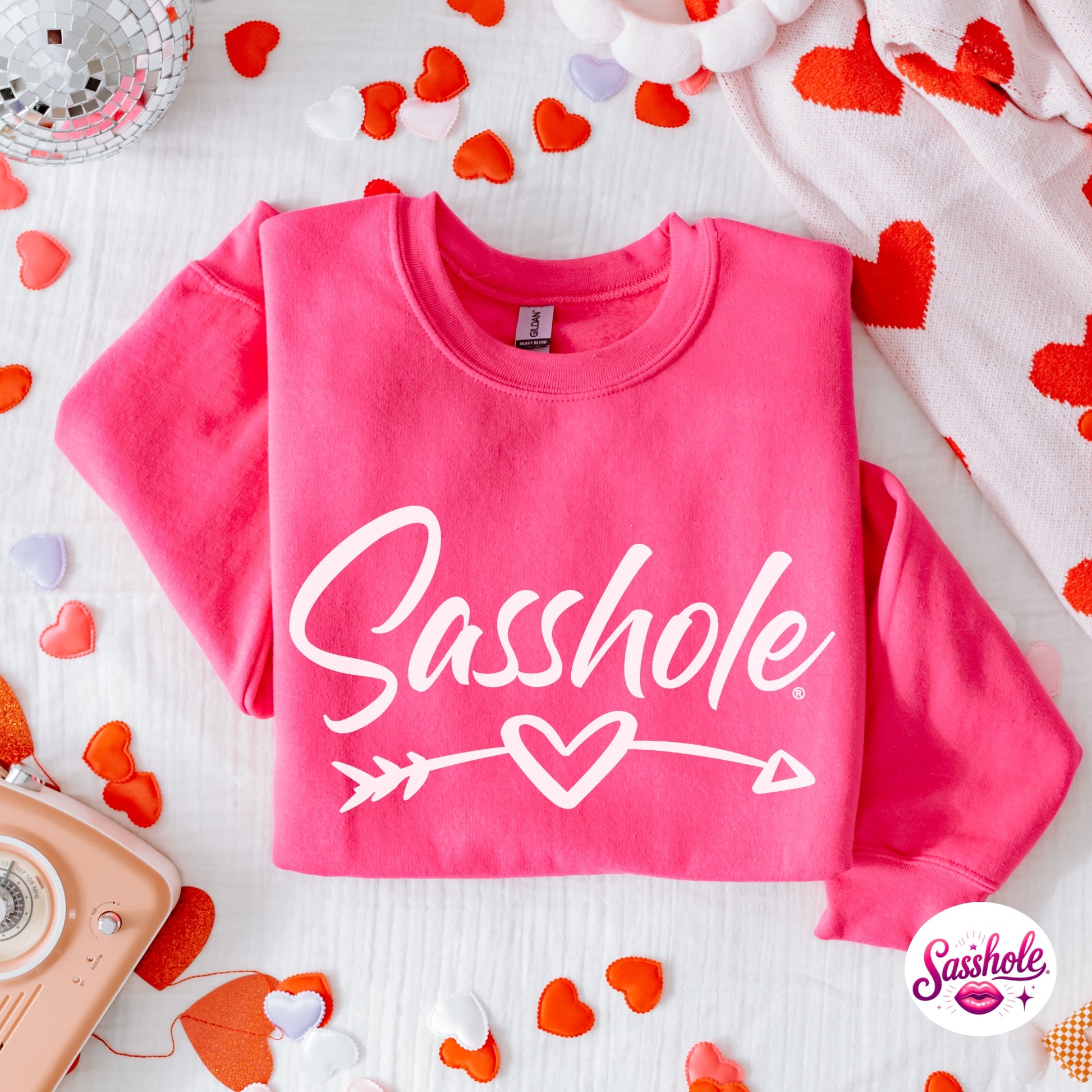 Pink sweatshirt with 'Sasshole' text surrounded by heart-shaped confetti on a light background