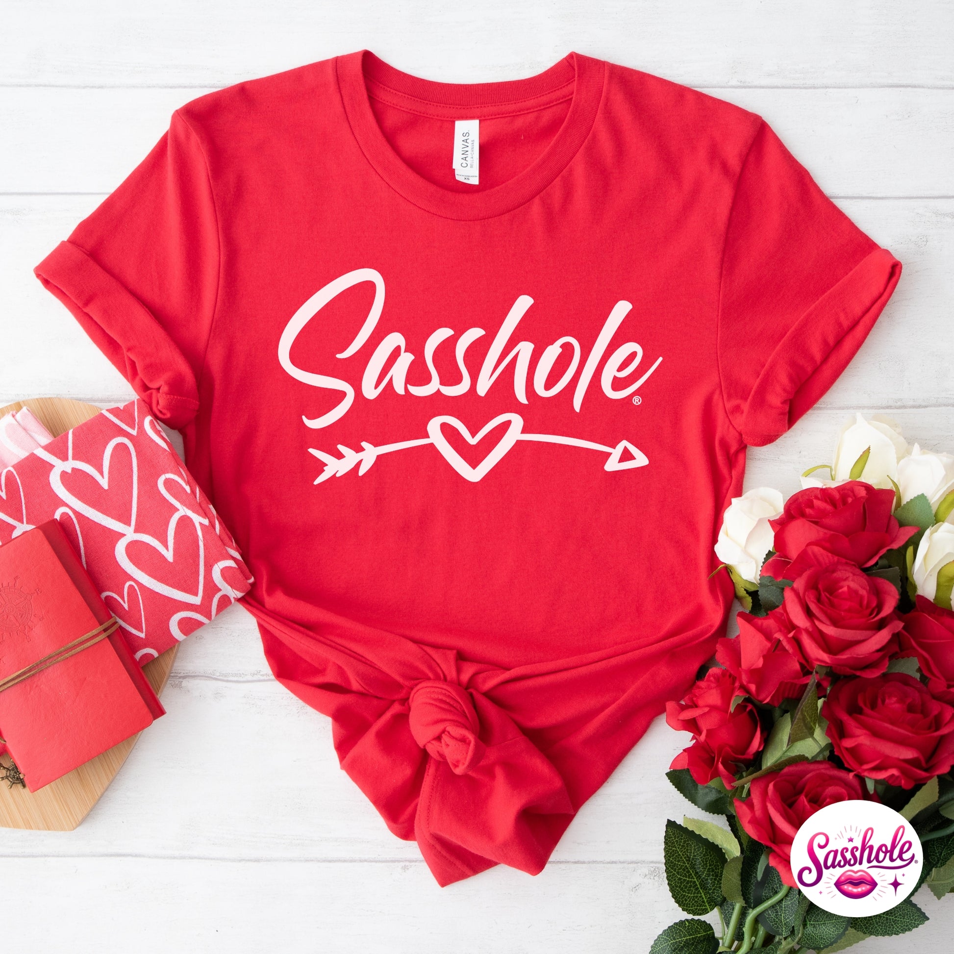 Red t-shirt with 'Sasshole' text on a white wooden surface with flowers and gift box.