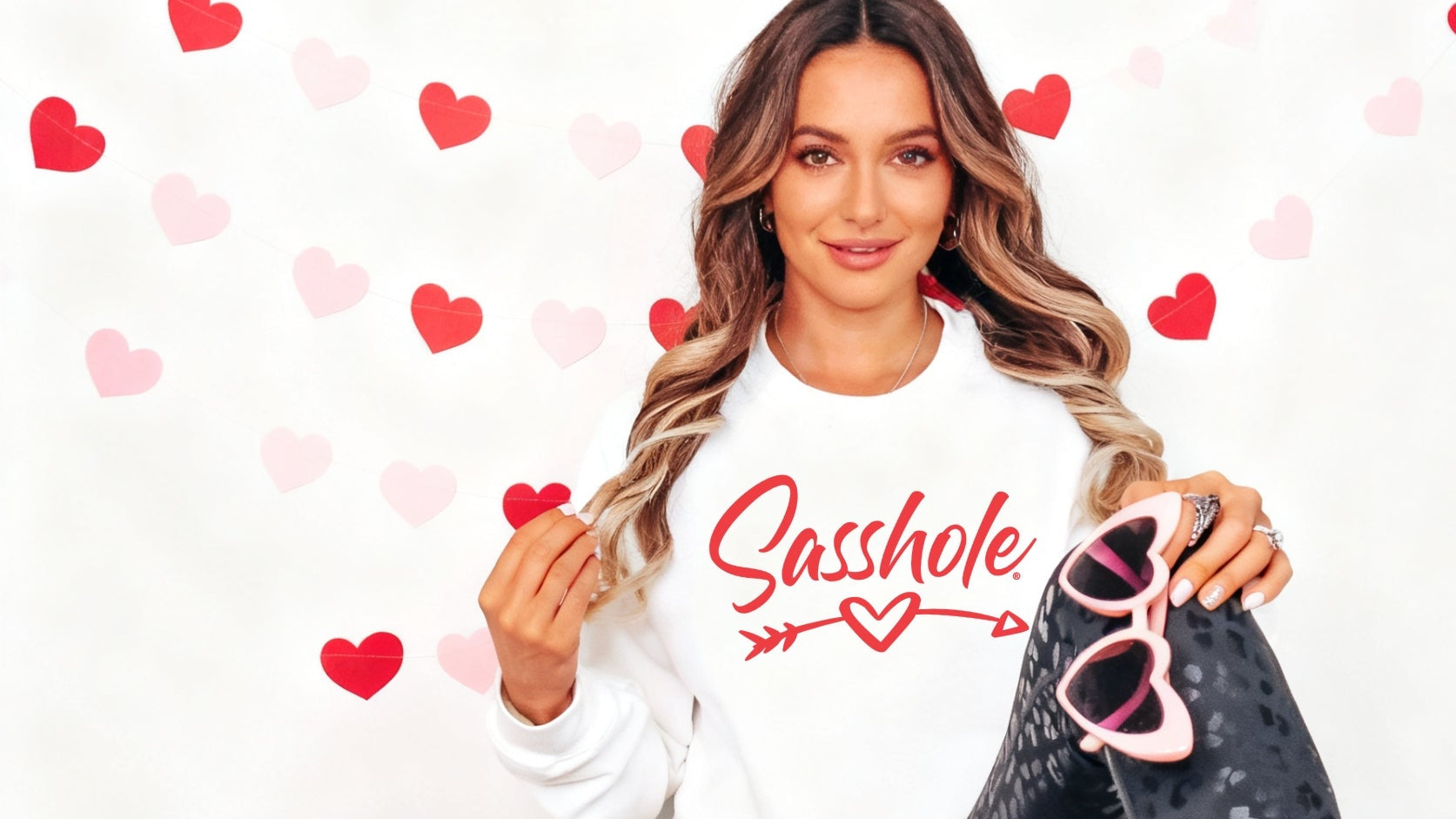 Woman wearing a white sweatshirt with 'Sasshole' printed on it, sitting on a white background with red hearts.