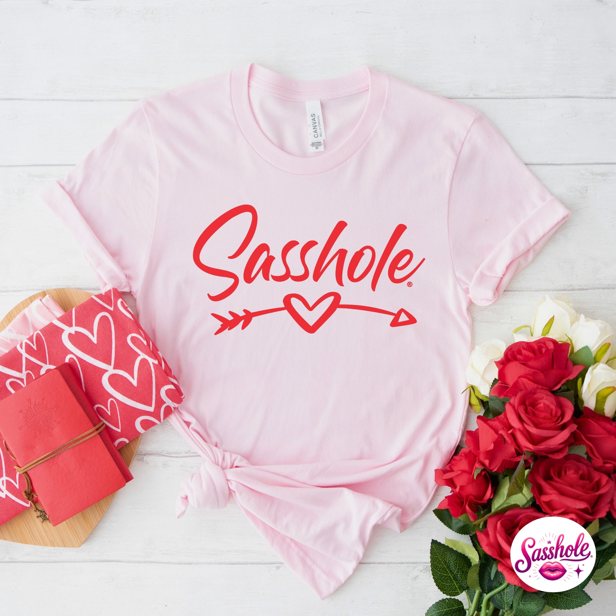 Image of Pink t-shirt with 'Sasshole' text, red roses, and gift box on a white wooden surface.