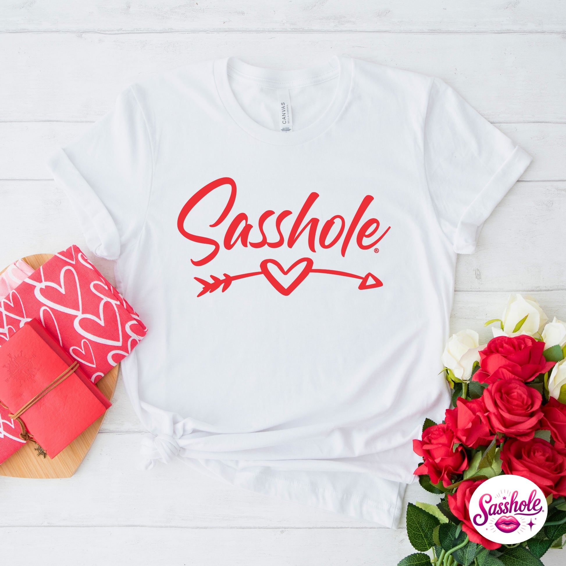 White t-shirt with 'Sasshole' text and red heart design on a white wooden surface with flowers and a pink box.